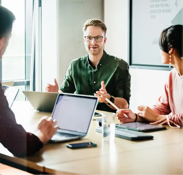 Three people sit at a conference table with laptops, having a discussion in a bright modern office.