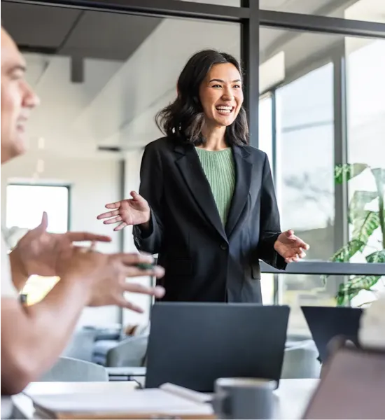 Smiling woman in a blazer stands and speaks to colleagues during a meeting in a bright, modern office.
