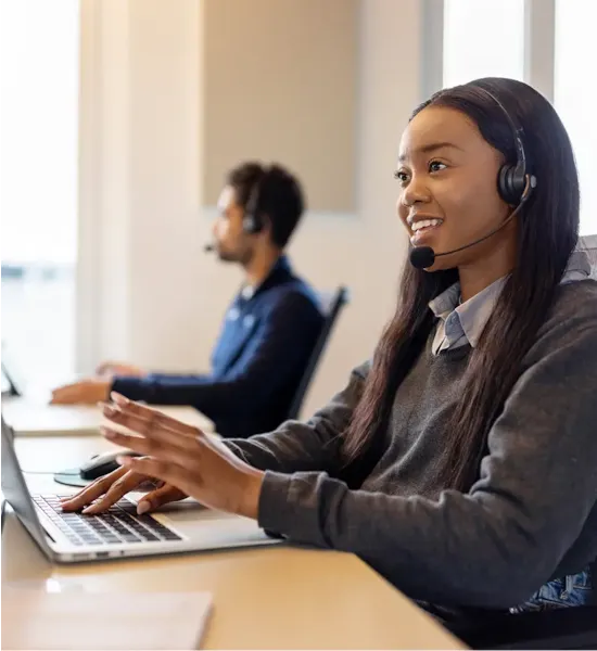 Smiling woman wearing a headset works on a laptop in an office, with a colleague in the background.